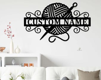 Pode incluir: Arte de parede em metal preto com uma bola de lã, uma agulha de tricô e o texto "CUSTOM NAME". O design inclui redemoinhos decorativos e é adequado para uma sala de artesanato ou como presente personalizado.