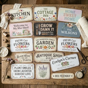 May include: A selection of decorative metal signs with a vintage aesthetic, featuring phrases like "Kitchen", "Home Sweet Home", and "Fresh Cut Flowers". The signs are adorned with floral motifs and garden-related themes.