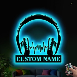 May include: A black headphone-shaped wall art piece with a blue neon glow. The design includes a sound wave graphic and a rectangular base with the text "CUSTOM NAME". A remote control is visible in the lower right corner.