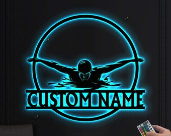 May include: A black metal wall art piece featuring a swimmer in a freestyle stroke, outlined with a bright blue neon glow. The text "CUSTOMNAME" is displayed below the swimmer. A remote control is visible in the lower right corner.