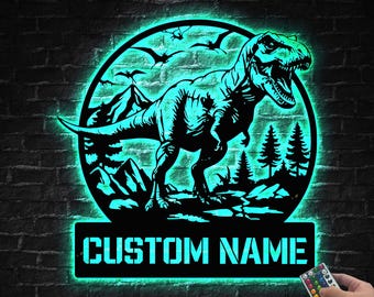 May include: A black metal sign featuring a Tyrannosaurus Rex silhouette within a circular frame, illuminated with a teal glow. The sign includes the text "CUSTOM NAME" below the dinosaur image, and is mounted on a brick wall.