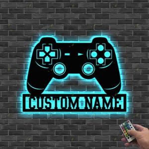 May include: Black illuminated wall art in the shape of a video game controller. The controller has a blue neon glow, with the text "CUSTOM NAME" below. A remote control is held in the lower right corner.