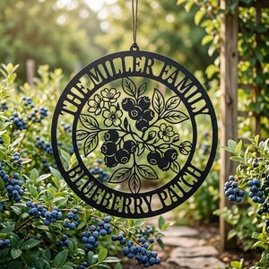 May include: A circular black metal sign featuring a cutout design of blueberry branches with leaves and blossoms. The text THE MILLER FAMILY is arched across the top, and BLUEBERRY PATCH is arched across the bottom, all suspended by a string.