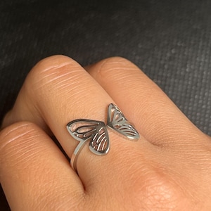Puede incluir: Un anillo plateado con un diseño de ala de mariposa. El anillo tiene un diseño abierto, con un ala a cada lado del dedo. Las alas de mariposa tienen un patrón calado detallado. El anillo se lleva en un dedo.