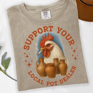 May include: Khaki-colored t-shirt with a graphic featuring a rooster smoking a cigarette, surrounded by the text "Support Your Local Pot Dealer." The design includes brown pottery and star accents. The shirt is a Comfort Colors brand.