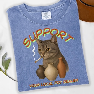 May include: Blue jean-colored t-shirt with a graphic of a cat smoking a cigarette, with the word "SUPPORT" above the cat and "YOUR LOCAL POT DEALER" below. Three small vases are in front of the cat.