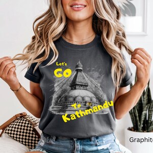 May include: Graphite-colored t-shirt with the text "Let's Go to Kathmandu" in yellow. The shirt features a graphic of a temple. The model is wearing blue jeans.