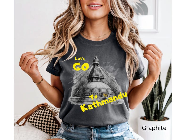 May include: Graphite-colored t-shirt with the text "Let's Go to Kathmandu" in yellow. The shirt features a graphic of a temple. The model is wearing blue jeans.