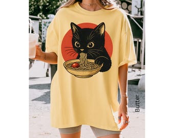 May include: Yellow oversized t-shirt featuring a black cat eating ramen from a bowl, set against a red circle. The cat illustration is detailed with yellow eyes and whiskers. The shirt has short sleeves and a relaxed fit.