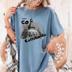 May include: A light blue t-shirt with the text "Let's Go to Kathmandu" and a black and white illustration of a temple. The shirt is made of a soft material.