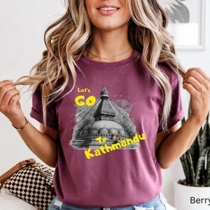 May include: Berry-colored t-shirt with the text "Let's Go to Kathmandu" in yellow. The graphic features a black and white image of a temple. The shirt is a short-sleeved crew neck.