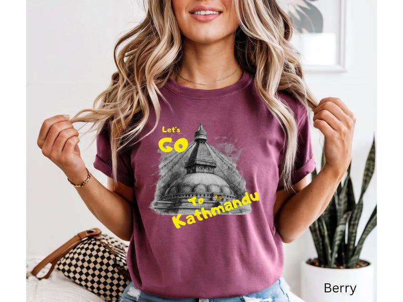 May include: Berry-colored t-shirt with the text "Let's Go to Kathmandu" in yellow. The graphic features a black and white image of a temple. The shirt is a short-sleeved crew neck.