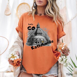 May include: Orange t-shirt with rolled-up sleeves featuring a graphic of a temple and the text "Let's Go To Kathmandu" in black. The shirt is a solid color and has a relaxed fit.