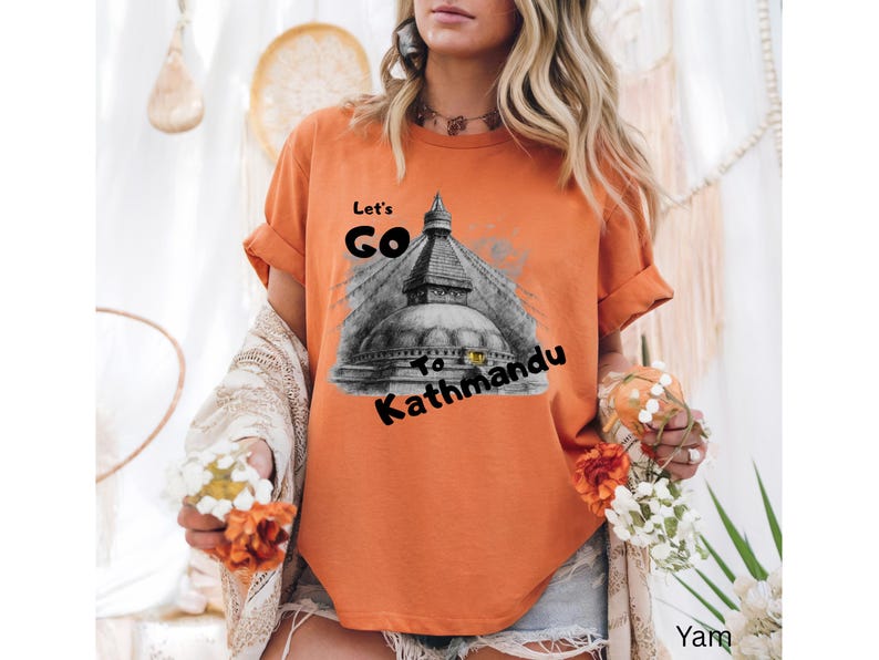 May include: Orange t-shirt with rolled-up sleeves featuring a graphic of a temple and the text "Let's Go To Kathmandu" in black. The shirt is a solid color and has a relaxed fit.