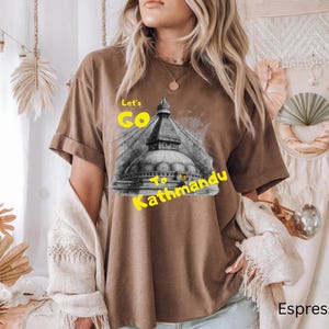 May include: Espresso-colored t-shirt featuring a graphic of a Nepalese stupa with the text "Let's Go to Kathmandu" in yellow. The shirt has rolled-up sleeves and a relaxed fit, suitable for casual wear.
