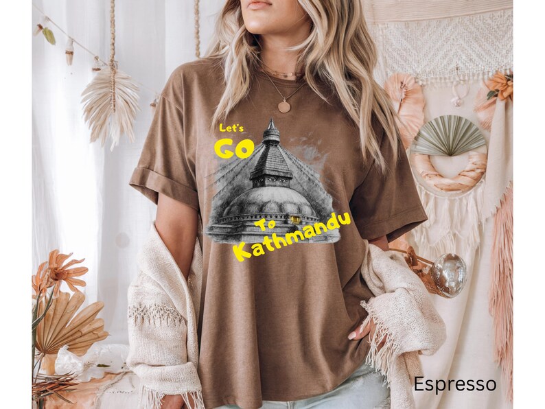 May include: Espresso-colored t-shirt featuring a graphic of a Nepalese stupa with the text "Let's Go to Kathmandu" in yellow. The shirt has rolled-up sleeves and a relaxed fit, suitable for casual wear.
