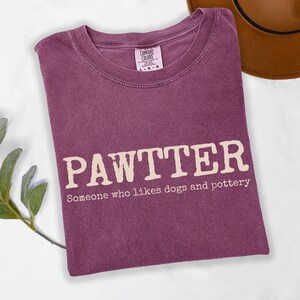 Pode incluir: Camiseta cor de baga com a palavra "PAWTTER" em negrito, em branco sujo. Abaixo da palavra, o texto diz "Algu&eacute;m que gosta de c&atilde;es e cer&acirc;mica". A camiseta est&aacute; dobrada, com um chap&eacute;u marrom vis&iacute;vel no fundo.