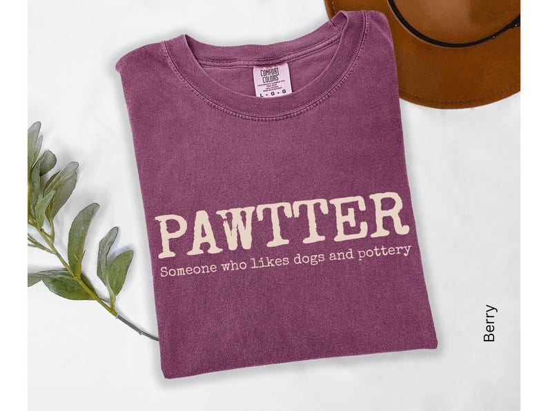 Pode incluir: Camiseta cor de baga com a palavra "PAWTTER" em negrito, em branco sujo. Abaixo da palavra, o texto diz "Algu&eacute;m que gosta de c&atilde;es e cer&acirc;mica". A camiseta est&aacute; dobrada, com um chap&eacute;u marrom vis&iacute;vel no fundo.
