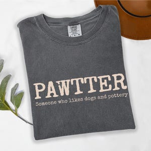 Pode incluir: Camiseta cinza escuro com a palavra "PAWTTER" em fonte marrom claro. Abaixo est&aacute; a frase "Someone who likes dogs and pottery". Um chap&eacute;u marrom &eacute; vis&iacute;vel ao fundo.