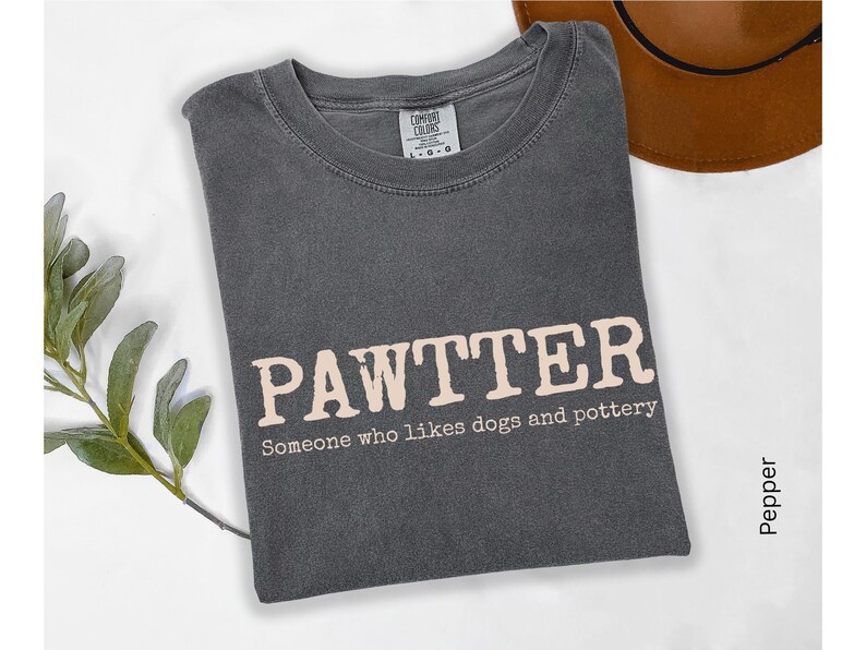 Pode incluir: Camiseta cinza escuro com a palavra "PAWTTER" em fonte marrom claro. Abaixo est&aacute; a frase "Someone who likes dogs and pottery". Um chap&eacute;u marrom &eacute; vis&iacute;vel ao fundo.