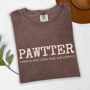 May include: Espresso-colored t-shirt with the word "PAWTTER" in large, off-white letters. Below, the text reads "Someone who likes dogs and pottery." The shirt is folded, with a brown hat visible in the background.