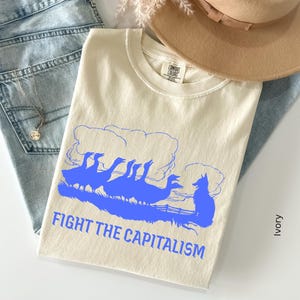 Pode incluir: Uma t-shirt cor de marfim com uma impressão gráfica azul de um grupo de gansos e uma raposa num campo com nuvens por cima. O texto "FIGHT THE CAPITALISM" está impresso em letras maiúsculas azuis abaixo do gráfico. Combinada com calças de ganga azuis.