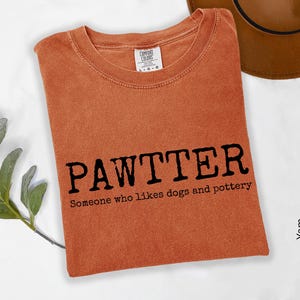 Pode incluir: Uma camiseta laranja dobrada com a palavra "PAWTTER" em preto e o texto "Someone who likes dogs and pottery" abaixo. Um chap&eacute;u marrom &eacute; vis&iacute;vel no fundo.