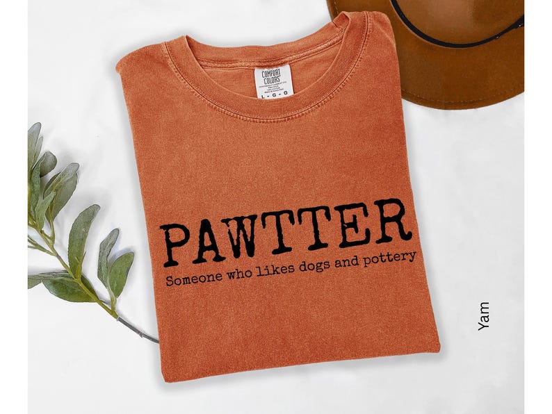 Pode incluir: Uma camiseta laranja dobrada com a palavra "PAWTTER" em preto e o texto "Someone who likes dogs and pottery" abaixo. Um chap&eacute;u marrom &eacute; vis&iacute;vel no fundo.