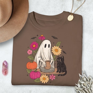 May include: Brown t-shirt featuring a whimsical illustration. A ghost is crafting pottery, surrounded by flowers, pumpkins, and a black cat. Bats are also included in the design. The design has a vintage, Halloween theme.