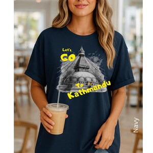 May include: Navy blue t-shirt with a graphic of a temple and the text "Let's Go to Kathmandu" in yellow. The image is of a woman holding a beverage.
