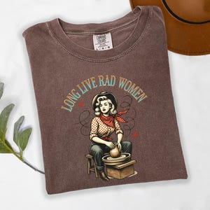 May include: Espresso-colored t-shirt with the text "LONG LIVE RAD WOMEN" and a graphic of a woman at a pottery wheel. The woman wears a cowboy hat, red bandana, and polka-dot shirt. The shirt is folded on a white surface.