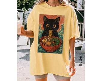 May include: Yellow t-shirt featuring a graphic of a black cat eating ramen from a red bowl. The cat is set against a backdrop of blue waves. The shirt is a casual, oversized fit.