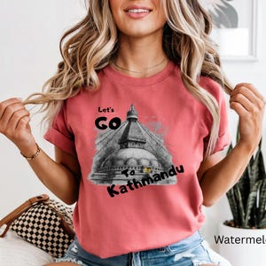 May include: A watermelon-colored t-shirt with the text "Let's Go To Kathmandu" in black. The graphic features a grayscale image of a temple. The shirt is a short-sleeved crew neck.