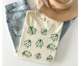 May include: Cream-colored t-shirt with a green ladybug pattern. The shirt is folded and placed on top of light blue jeans, with a tan hat and a necklace visible in the background. The shirt has a tag that says "COMFORT COLORS".