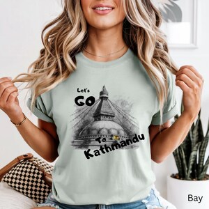 May include: A light green t-shirt with the text "Let's Go To Kathmandu" and a black and white illustration of a temple. The shirt is a short-sleeved crew neck.