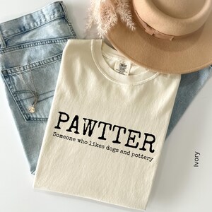 Pode incluir: Camiseta cor marfim com a palavra "PAWTTER" em preto e a frase "Someone who likes dogs and pottery". A camiseta est&aacute; sobre jeans azul claro, com um chap&eacute;u bege e raminhos decorativos.