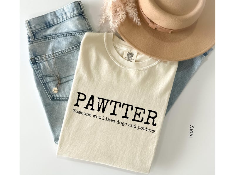 Pode incluir: Camiseta cor marfim com a palavra "PAWTTER" em preto e a frase "Someone who likes dogs and pottery". A camiseta est&aacute; sobre jeans azul claro, com um chap&eacute;u bege e raminhos decorativos.