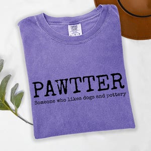 Pode incluir: Camiseta violeta com a palavra "PAWTTER" em letras pretas em negrito e a frase "Someone who likes dogs and pottery" abaixo. Um chap&eacute;u marrom &eacute; vis&iacute;vel ao fundo.