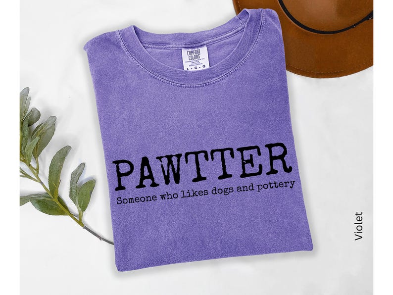 Pode incluir: Camiseta violeta com a palavra "PAWTTER" em letras pretas em negrito e a frase "Someone who likes dogs and pottery" abaixo. Um chap&eacute;u marrom &eacute; vis&iacute;vel ao fundo.