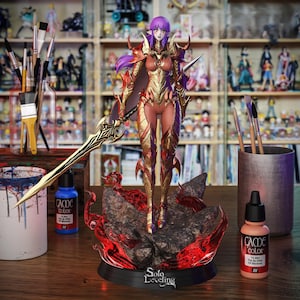 May include: A detailed figurine of a female character from "Solo Leveling" stands on a rocky base with fiery accents. She wears gold and red armor, holds a large sword, and has purple hair. Paint supplies and other figurines are in the background.