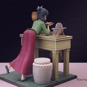 May include: A figurine of a woman in a long red skirt and green top, leaning on a beige table. A grinding wheel and a jar with a lid are next to the table. The figurine is on a green base.