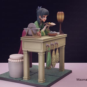 May include: A detailed figurine of a woman in a green outfit, seated at a table. The table has a lamp, a basket, and other items. The figurine is on a green base with a pink background. The figurine is labeled "Maomao".
