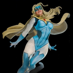 Op de afbeelding: Een superheldenfiguur met een blond personage in een blauw-wit turnpakje met bijpassend masker. Het figuur draagt een gouden cape met een rood ster-embleem bij de nek, afgebeeld in een dynamische vlieghouding met lang wapperend haar.