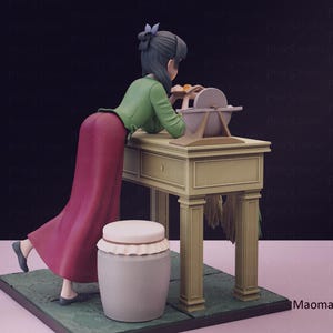 May include: A figurine of a woman in a long red skirt and green jacket, leaning over a table with a grinding wheel. A white jar with a lid sits nearby. The scene is set on a green base.