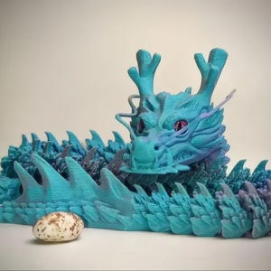 May include: A 3D-printed articulated dragon in shades of teal and purple, with a detailed head and spiky back. The dragon is posed on a white surface, with a speckled egg nearby. The dragon's eyes are pink.