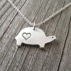 May include: A silver-colored turtle pendant necklace. The turtle has a heart-shaped cutout on its shell and a small dot for an eye. The necklace chain is a delicate silver color.