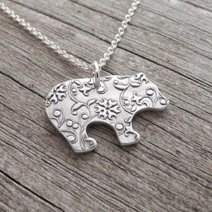 A silver-colored pendant in the shape of a bear with a floral design on it, hanging from a chain.