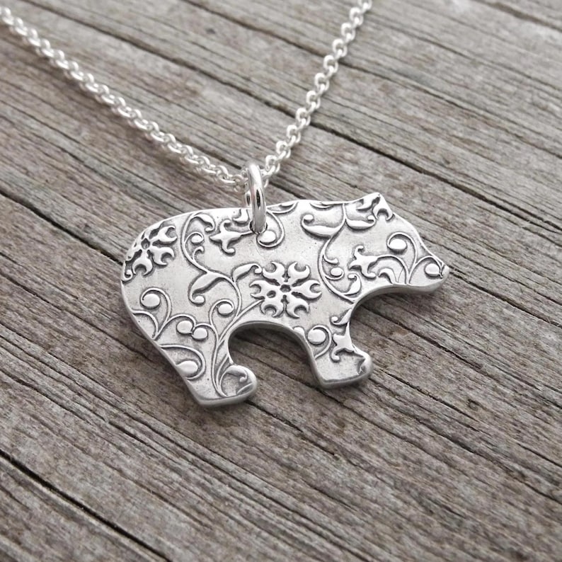 A silver-colored pendant in the shape of a bear with a floral design on it, hanging from a chain.
