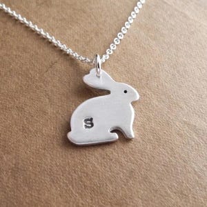 May include: A silver-colored bunny pendant necklace. The bunny is in a sitting position with a small black dot for an eye and the letter "S" engraved on its side. The necklace chain is also silver.
