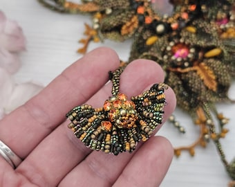 May include: A beaded pendant in shades of green, black, and gold, with orange accents. The pendant is fan-shaped and features intricate beadwork. The background shows a larger, similar beaded piece. Jewelry.